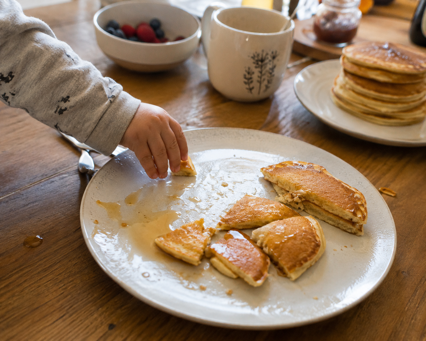 A relaxed breakfast table with pancakes, toddler's hands reaching for food, casual weekend mood