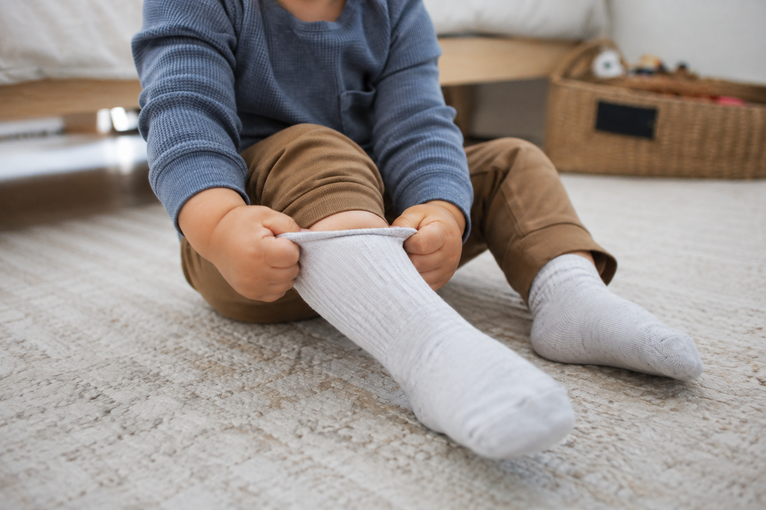 Toddler's hands pulling on a sock unassisted, slightly crooked, sitting on bedroom floor